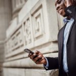 Low angle de profil d'un homme afro-américain positif en costume élégant utilisant des écouteurs sans fil tout en ayant une conversation téléphonique près d'un bâtiment contemporain