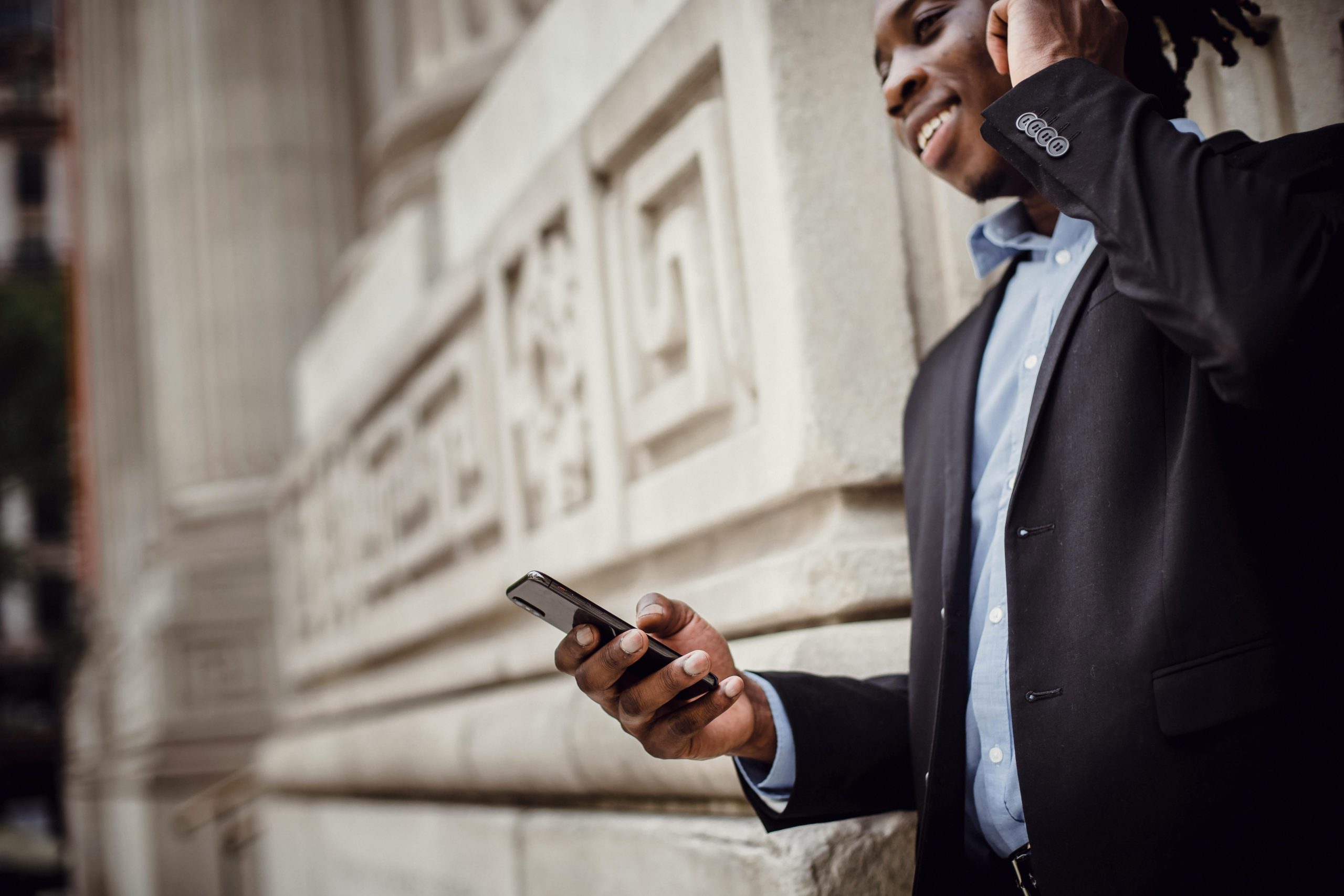 Low angle de profil d'un homme afro-américain positif en costume élégant utilisant des écouteurs sans fil tout en ayant une conversation téléphonique près d'un bâtiment contemporain