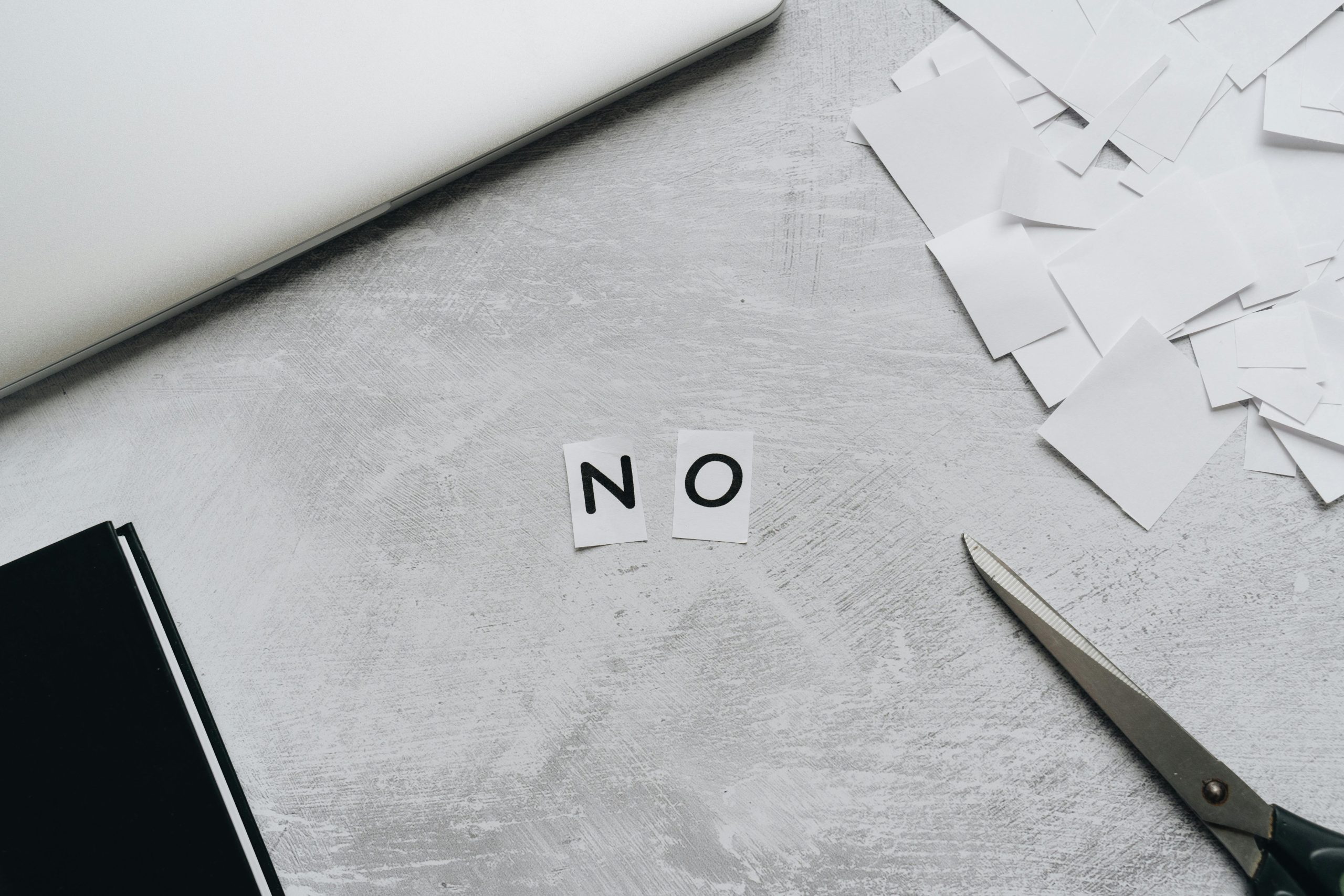 Top view of scattered paper squares, laptop, and scissors forming the word 'NO', implying rejection or denial. Vue de dessus de carrés de papier dispersés, d'un ordinateur portable et de ciseaux formant le mot « NON », impliquant un rejet ou un refus.