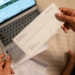Individual holding a cheque over laptop, signifying online banking or financial transaction. Un individu tenant un chèque au-dessus d'un ordinateur portable, signifiant les services bancaires en ligne ou une transaction financière.