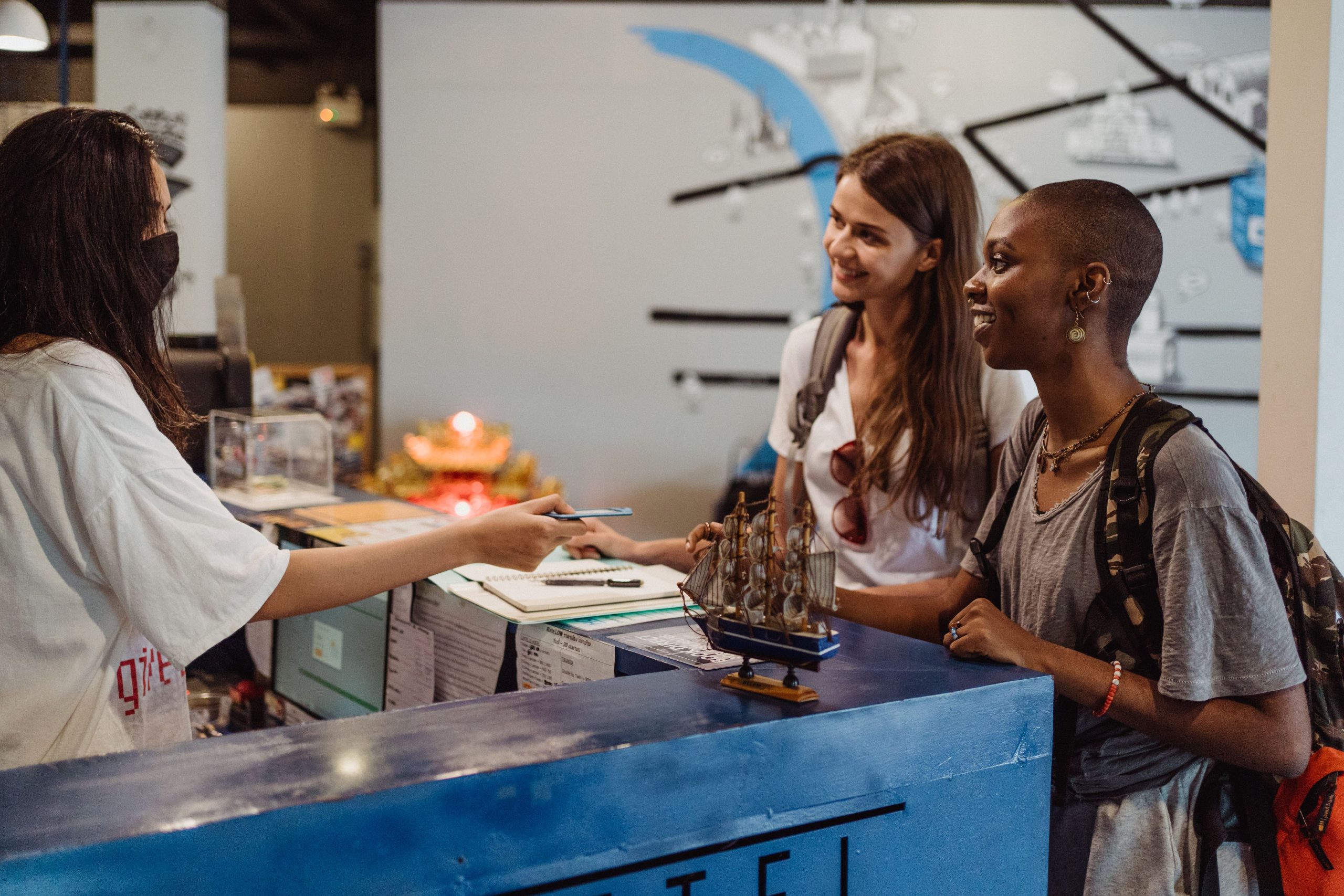 Two women travelers checking in at a hostel reception with a welcoming staff member wearing a face mask. Deux femmes voyageurs effectuant l'enregistrement à la réception d'une auberge avec un membre du personnel accueillant portant un masque facial.